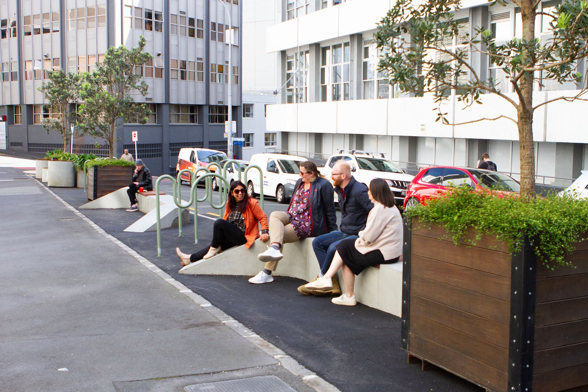 A diverse group of five people sit on the concrete seating amongst the planter boxes and bike racks at St Paul street in downtown Auckland. The trees are hemmed by native climbing plants.