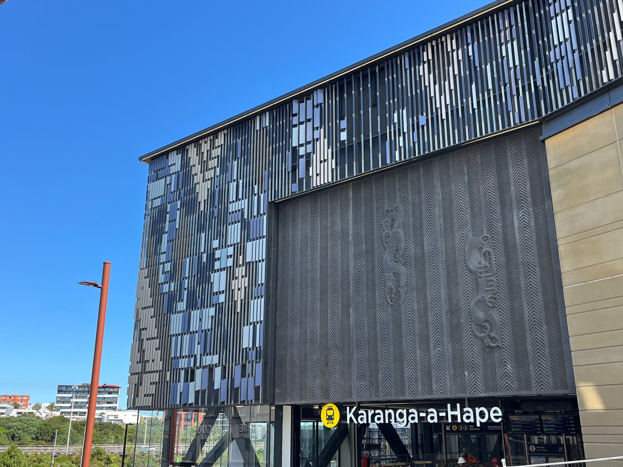 The upper portion of Karanga-a-Hape rail station building, including the station name signage; the threshold element above the entrance with two moulded manaia (guardian figures) placed on a base reflecting traditional angular carving; surrounded by angled metal fins creating a pattern reflective of Te Ika-whenua-o-te-rangi, the Milky Way constellation. 
