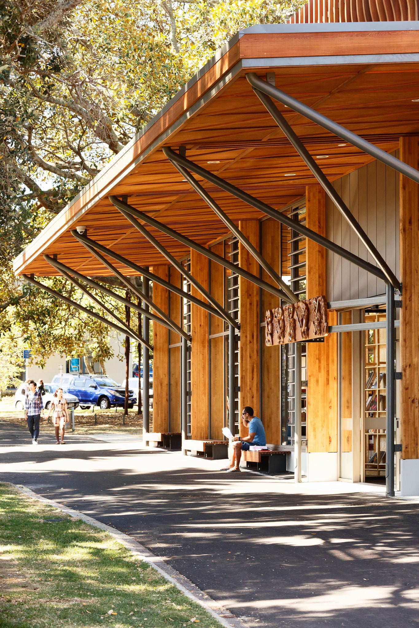 A man is sitting outside the library, partly in sun and partly in shade under the wooden awning.