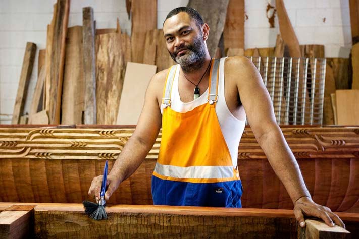 Local artist: Whare Thompson. He is a māori man with a beard, holding a paintbrush and in work clothes poses proudly in front ol wooden carvings in his workshop.f intricate, beautifu