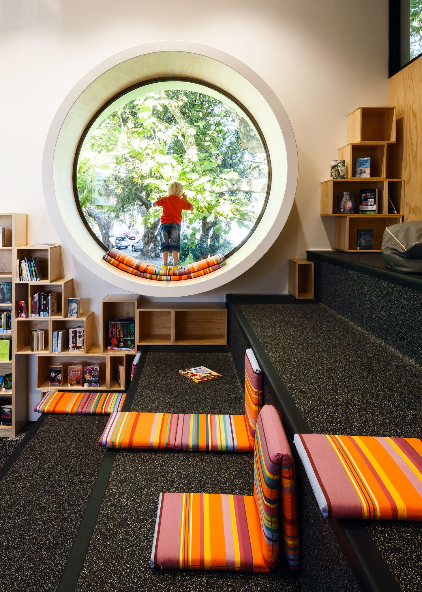 A young child is alone, looking out of the large port hole window over to the playground across the street.