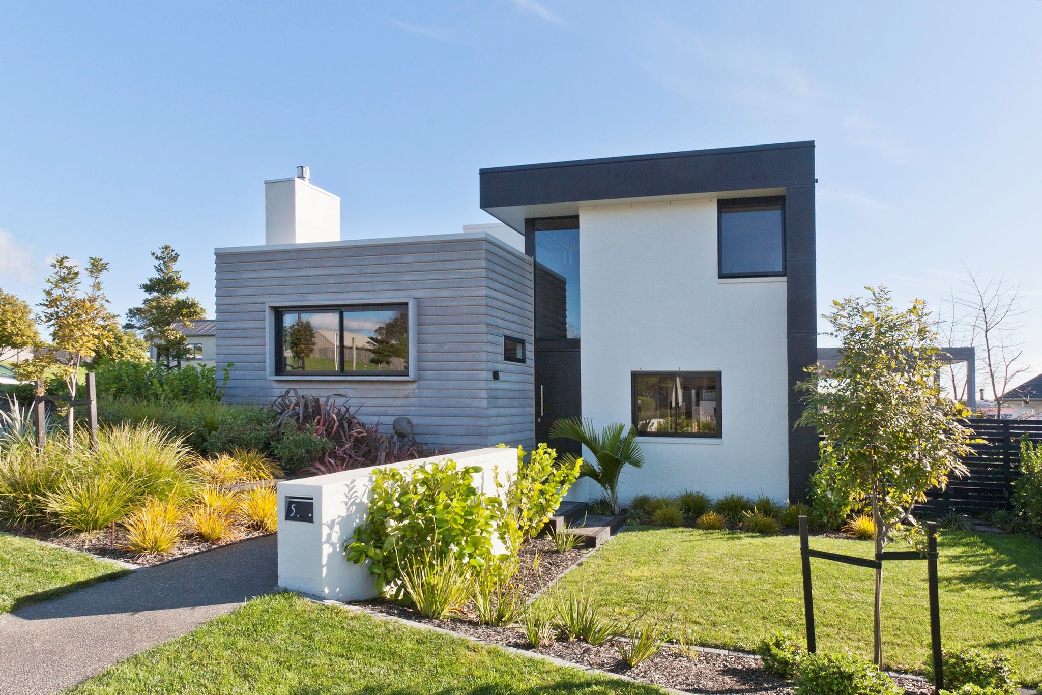 Photo of a single level and attached 2 storey space which combined is the house, facing the road surrounded by a garden.