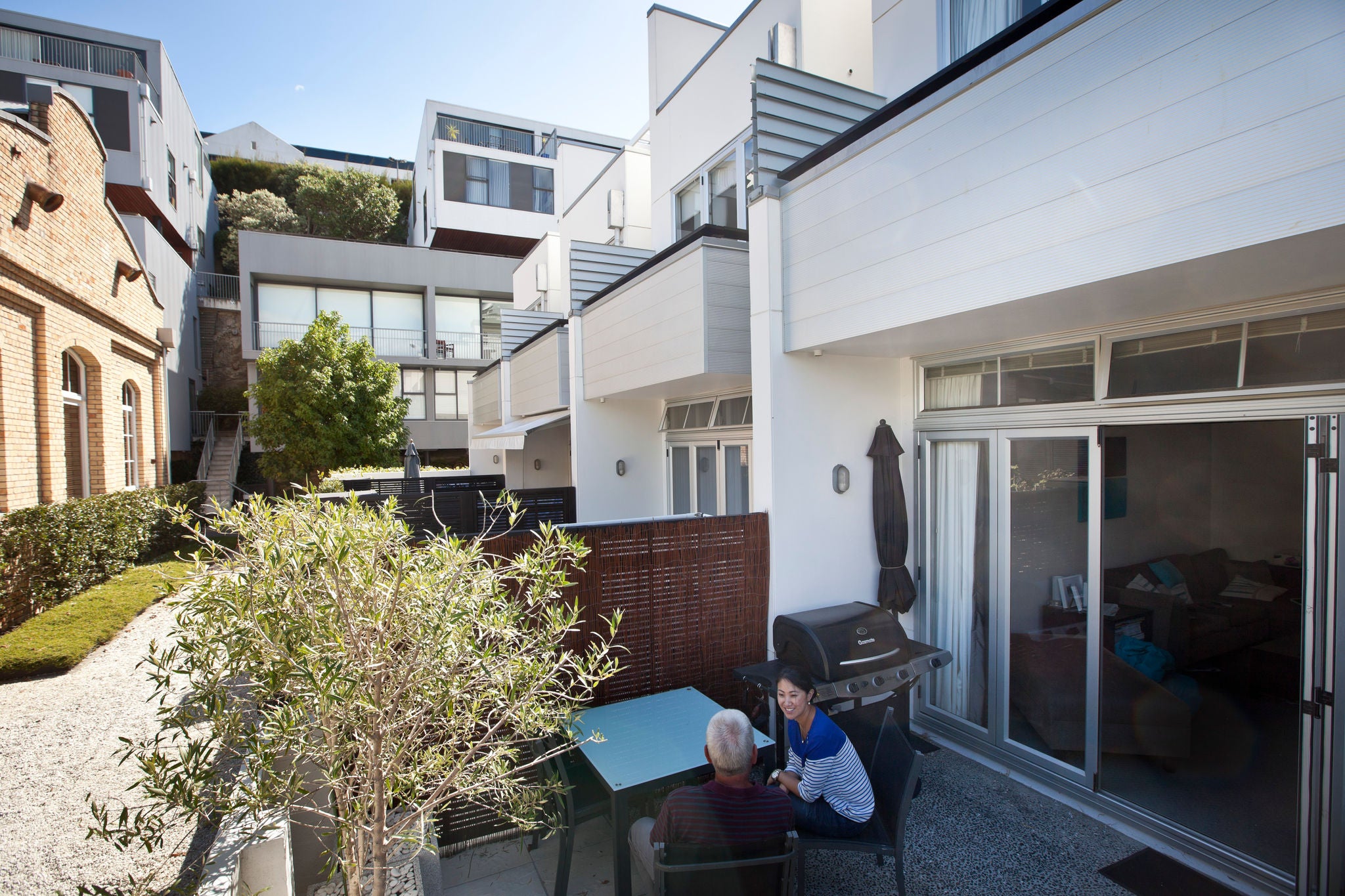 A shaded and cosy backyard with plants and a BBQ. People are seated outside having a chat.