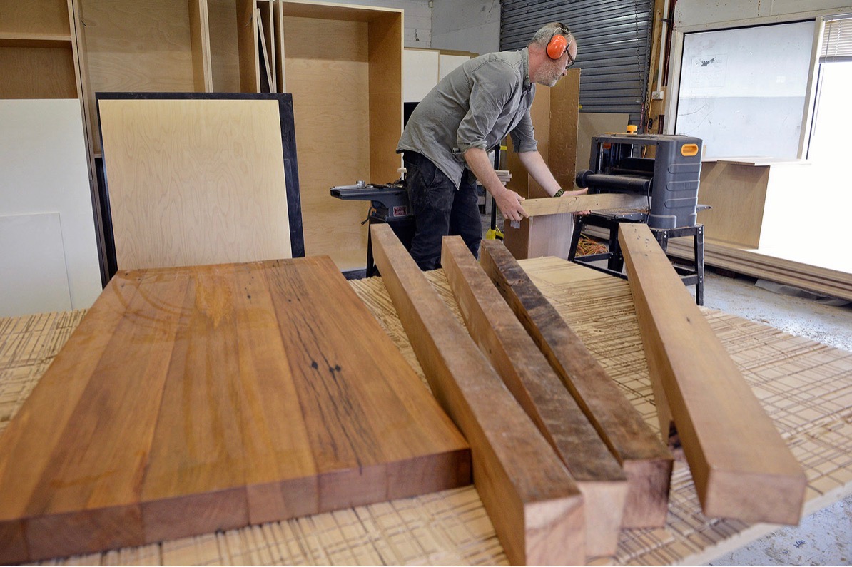 A carpenter in a workshop, feeding the rimu beams into a mechanical planer.