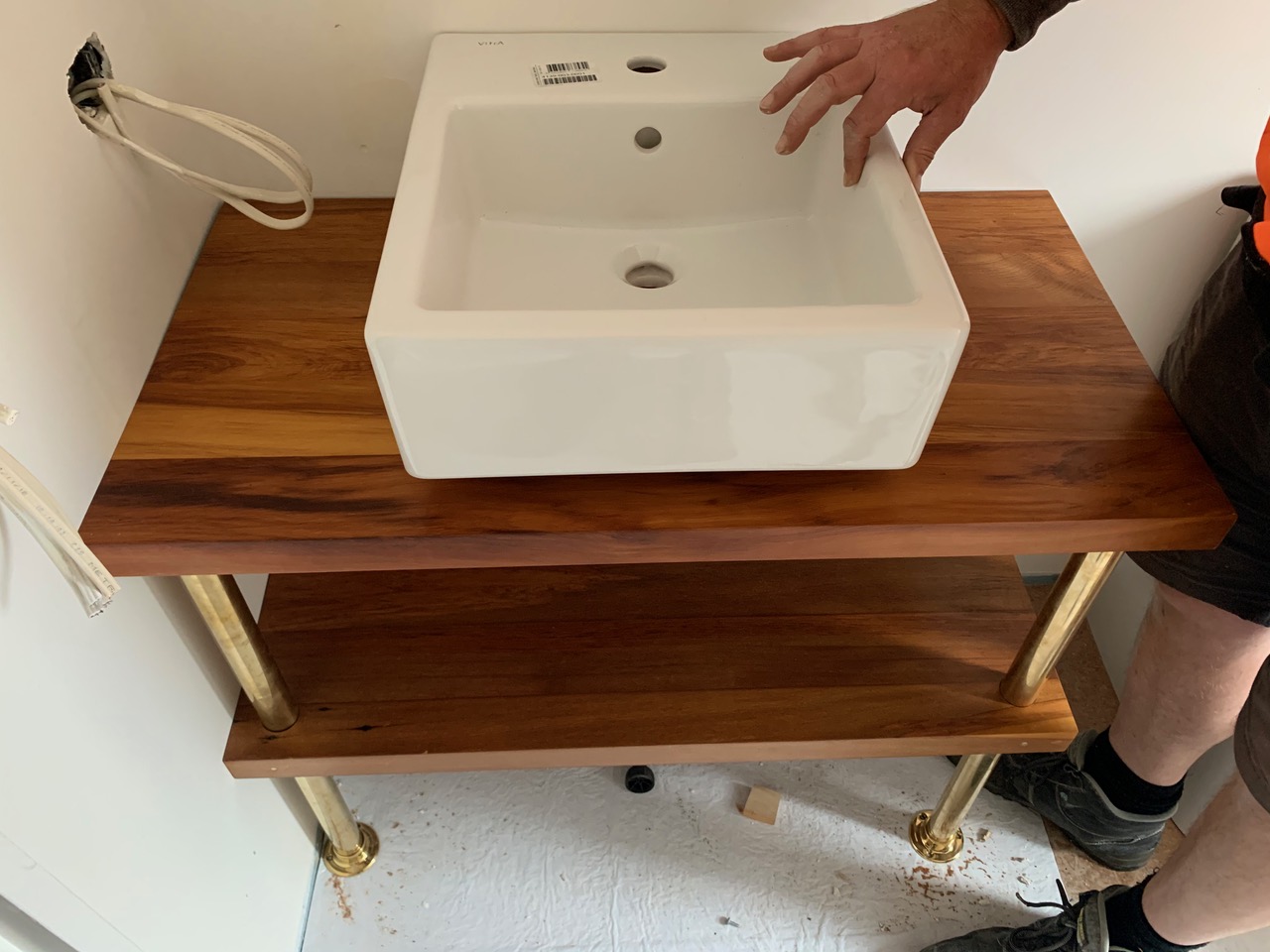 A plumber places a new bathroom basin on a plinth made out of two rimu boards in a small bathroom.