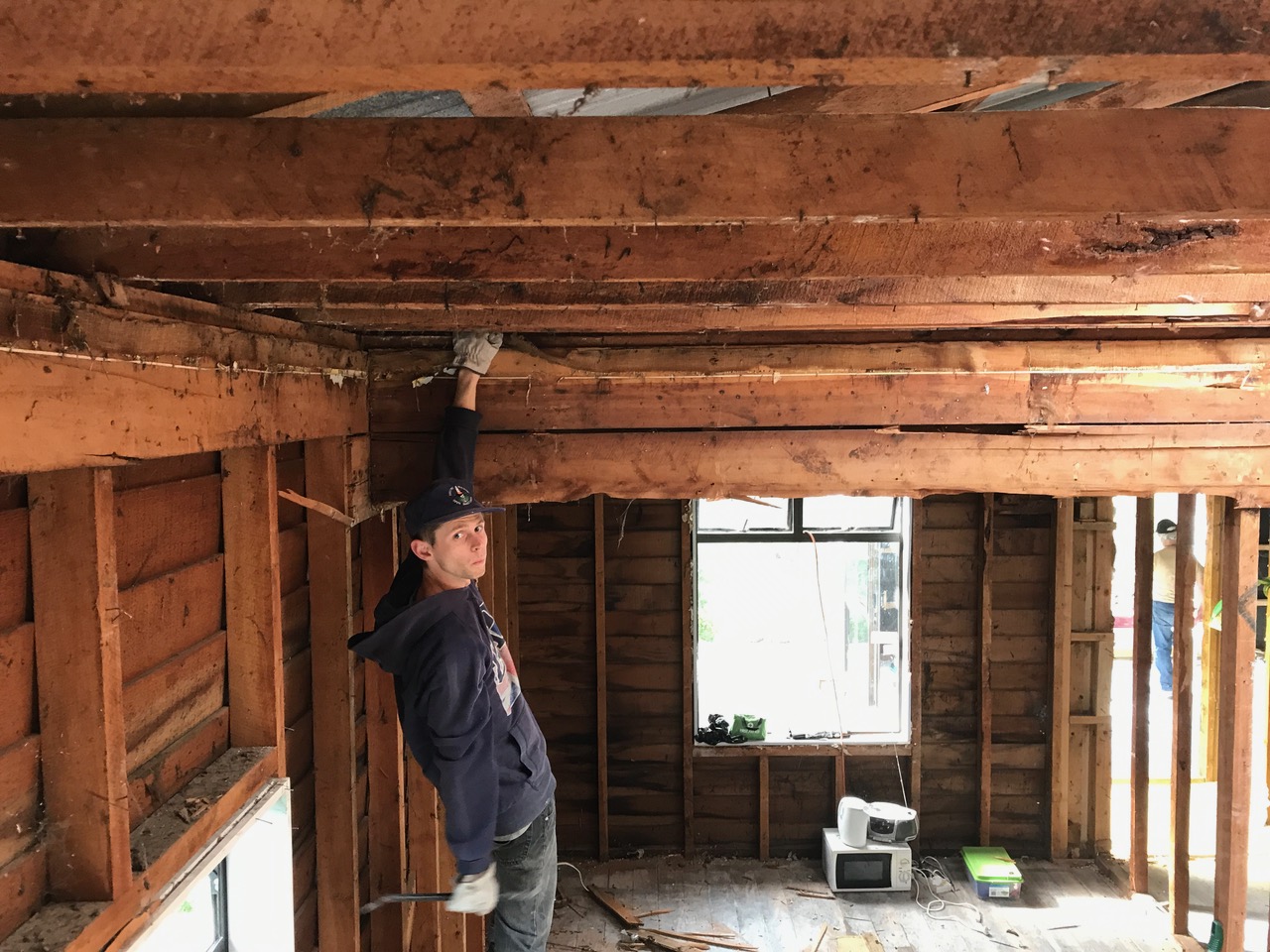 A young man wearing gloves and holding a crowbar is pulling beams out of the bungalow's ceiling.