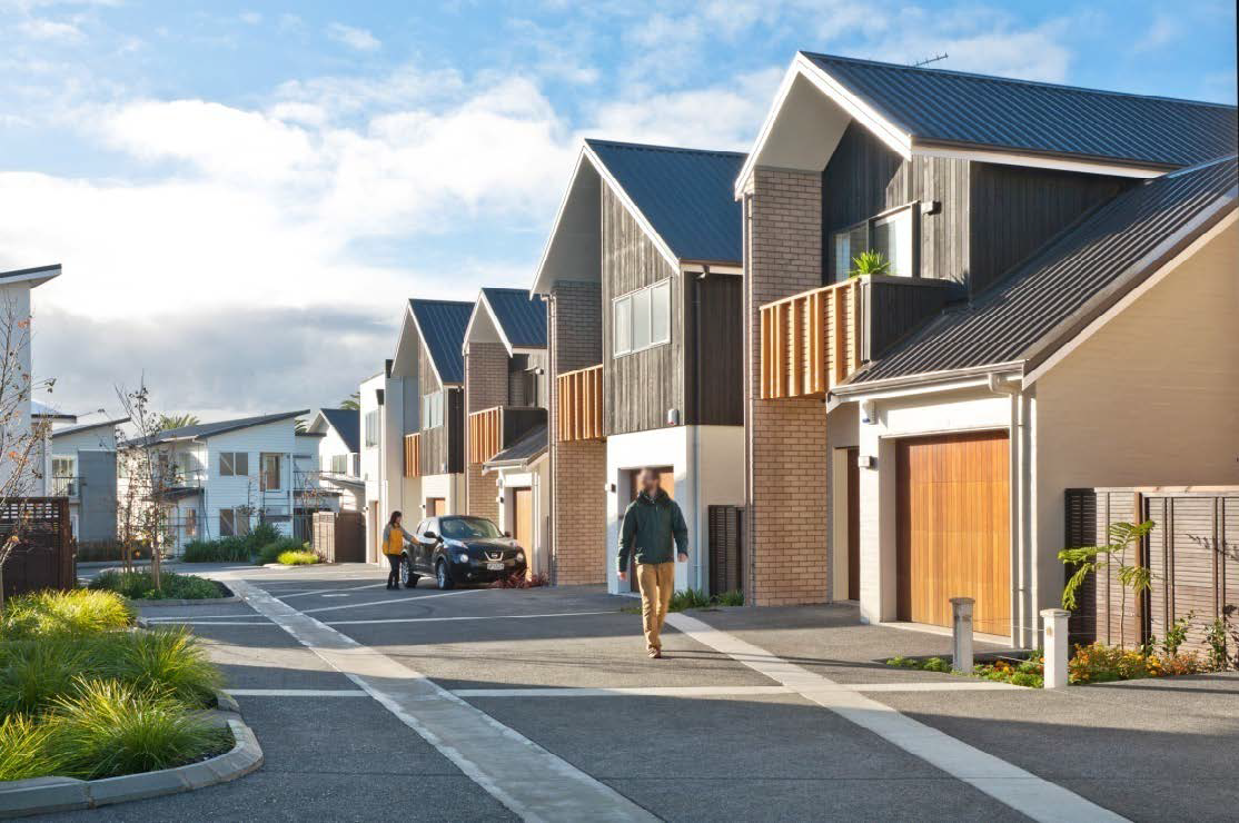 Looking at the homes from the street in the evening light; there are people walking around.