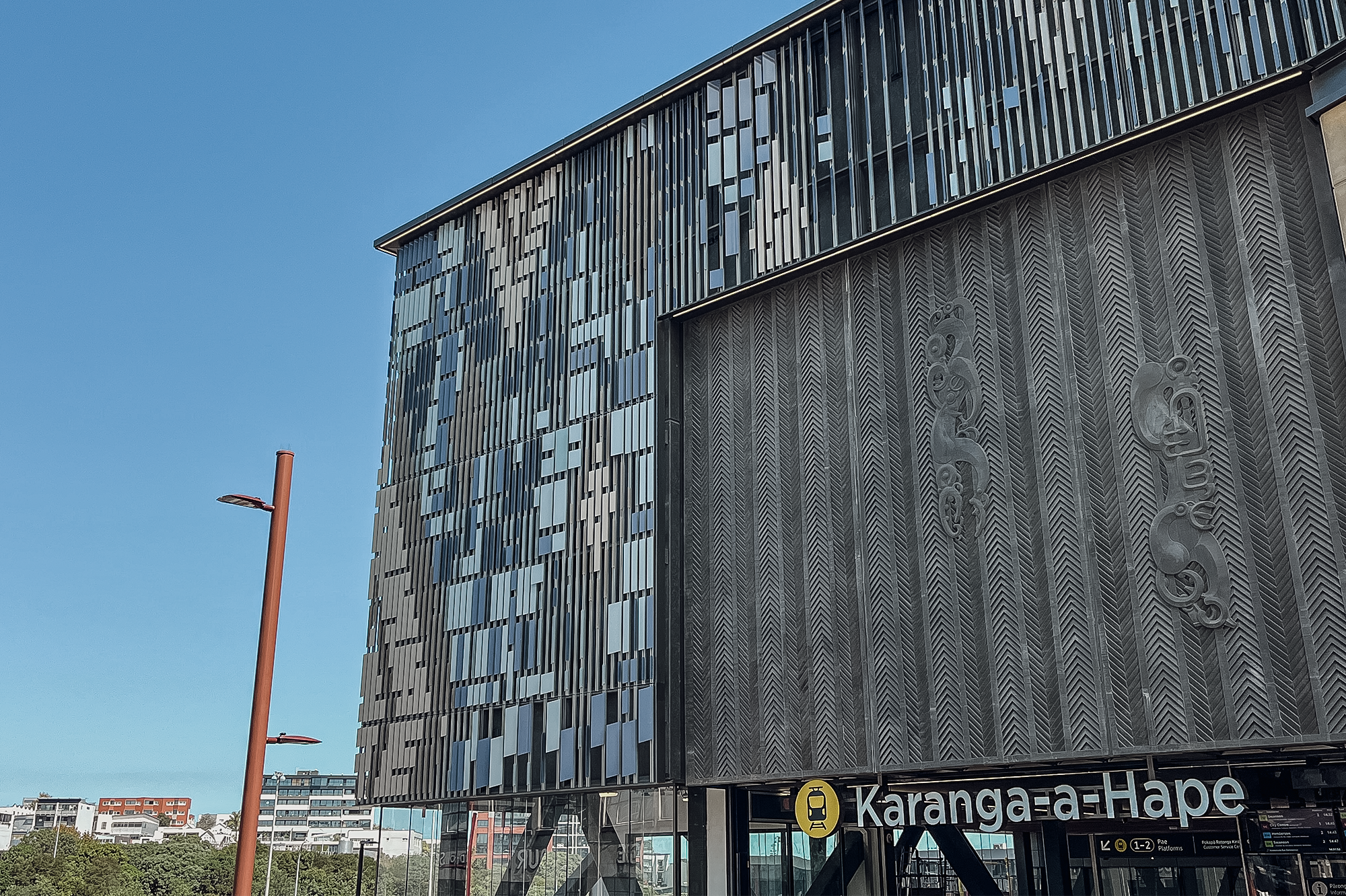 The upper portion of Karanga-a-Hape rail station building, including the station name signage; the threshold element above the entrance with two moulded manaia (guardian figures) placed on a base reflecting traditional angular carving; surrounded by angled metal fins creating a pattern reflective of Te Ika-whenua-o-te-rangi, the Milky Way constellation.