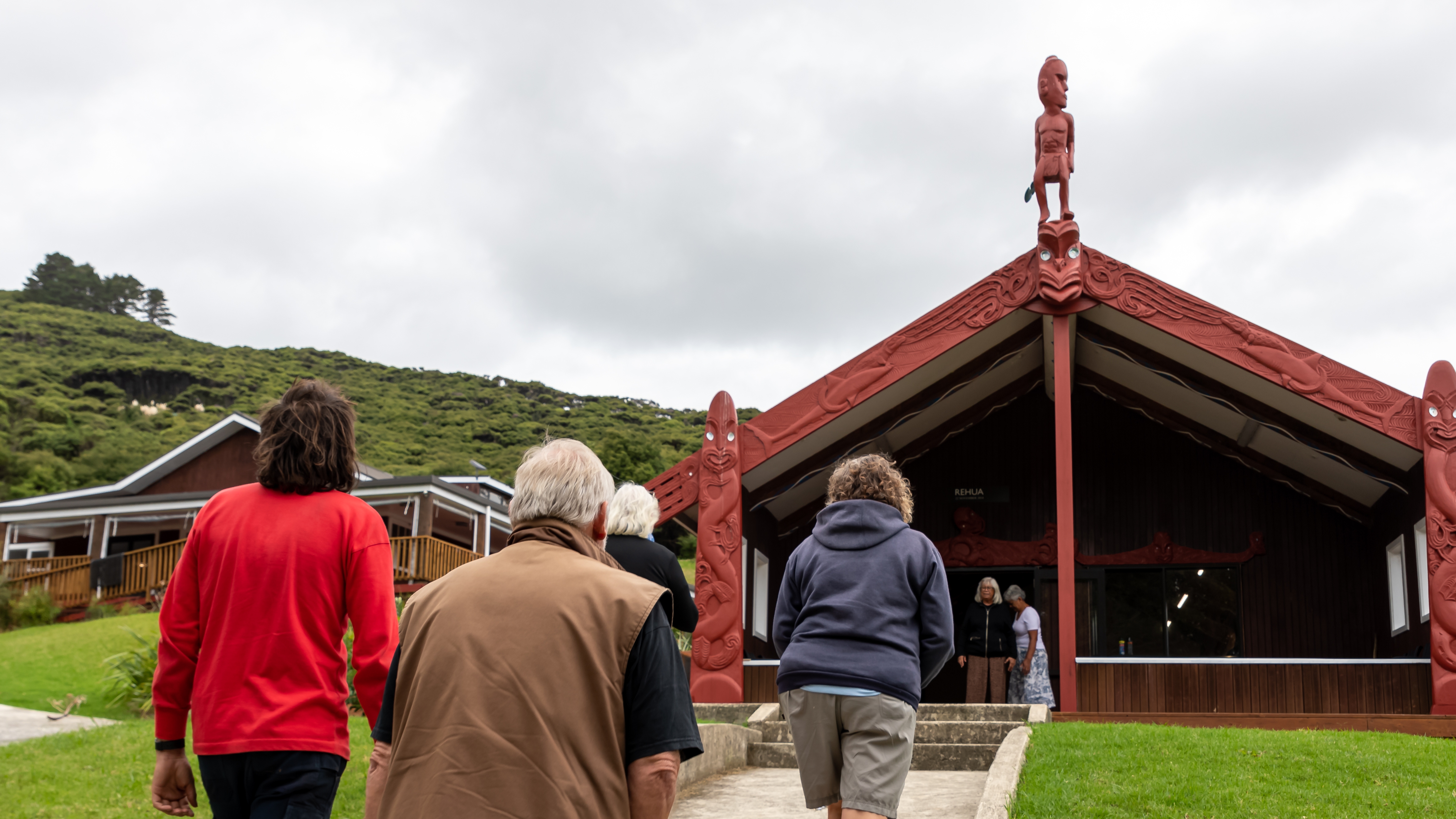 Four people approaching the wharenui of Kawa Marae, Aotea Great Barrier Island. 